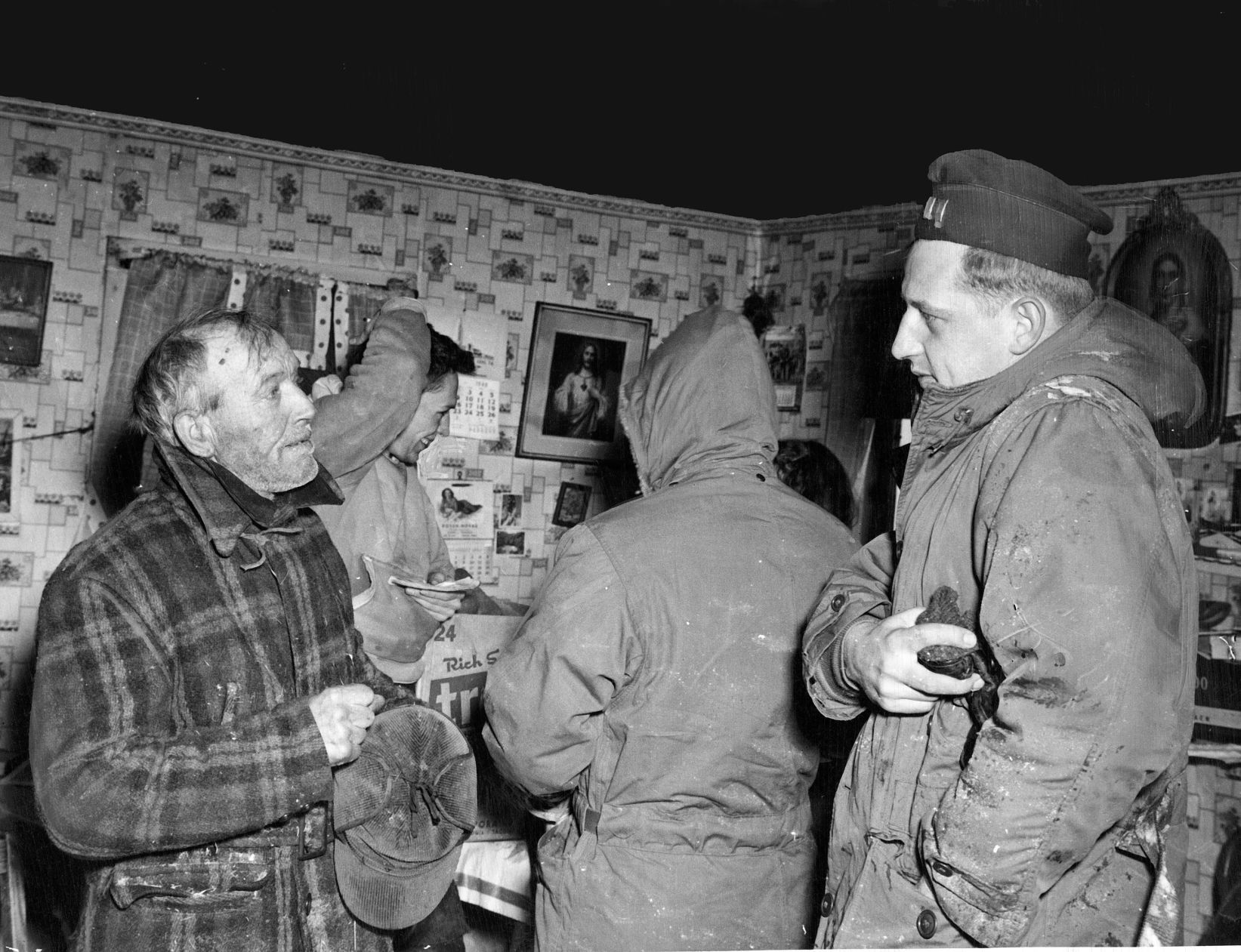 At the Carl elder farm, 8 miles from O'Neill, a Fifth Army relief group traveling by weasel found the family in a bad plight. The Belzers were low on food and had been burning brush to supplement their meager fuel supply. Mr. Belzer (left) is shown chatting with Capt. William Tanski (right.) World-Herald reporter Paul Williams is interviewing Mary Belzer (hidden behind unidentified soldier.) Published Feb. 4, 1949.